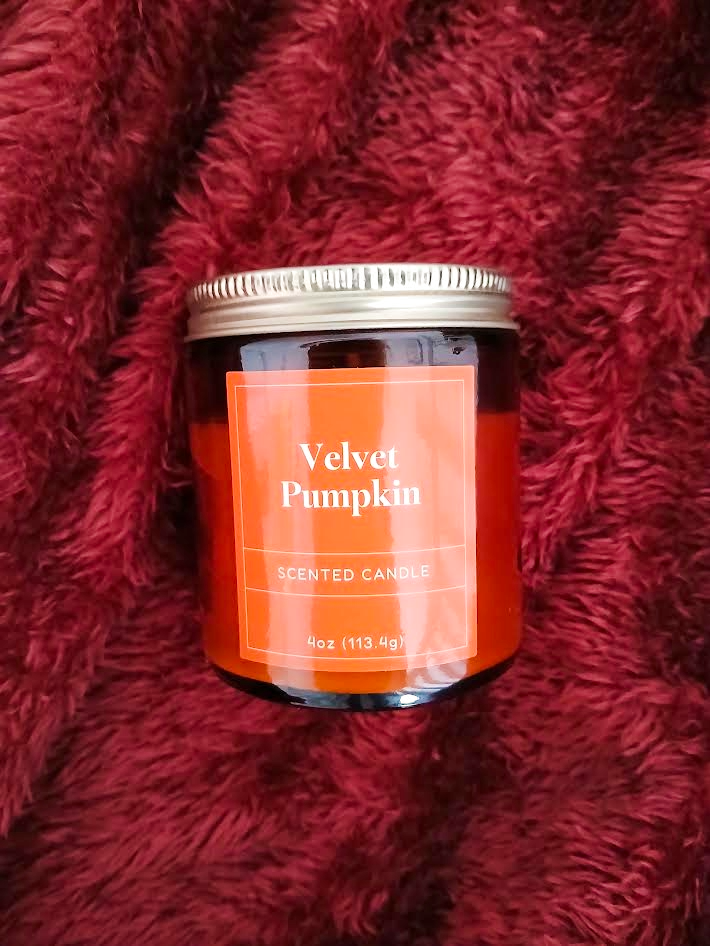 A 4oz scented candle jar labeled 'Velvet Pumpkin' resting on a deep red fluffy surface.