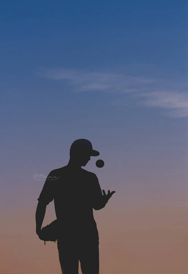 Silhouette of a person holding a baseball and tossing it against a sunset background.
